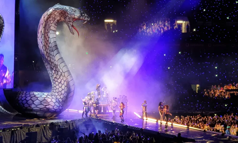a stage in a packed stadium with Taylor Swift performing in the centre, dancers around her and a large silver snake prop to the left
