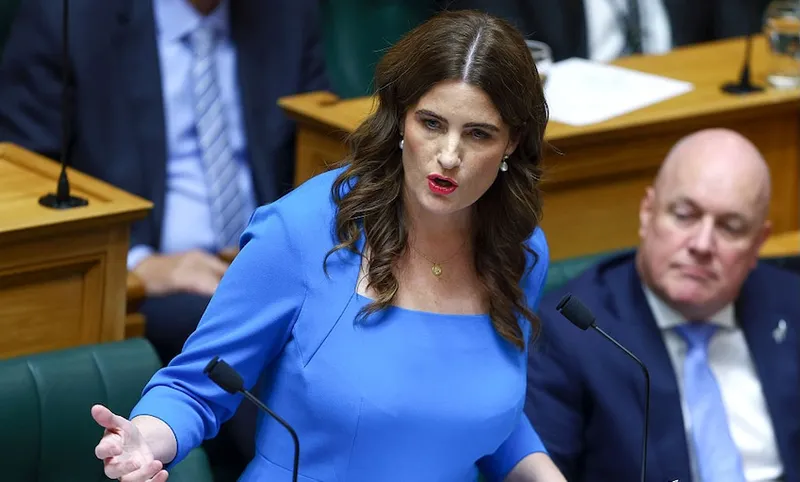 A woman in a blue dress speaks passionately at a podium in a government chamber, while a man in a suit sits and listens behind her.