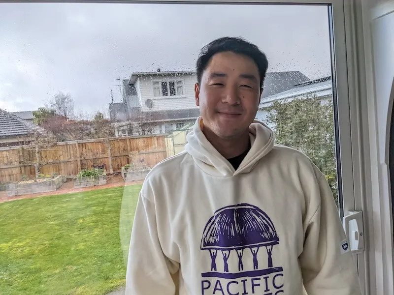 a young man in a pacific studies hoody, smiling at the camera with a rainy garden outside