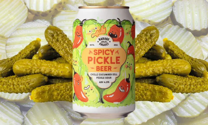 A can of Spicy Pickle Beer with a colorful label featuring cartoon pickles and peppers is placed in front of a pile of pickles, with sliced pickles in the background.