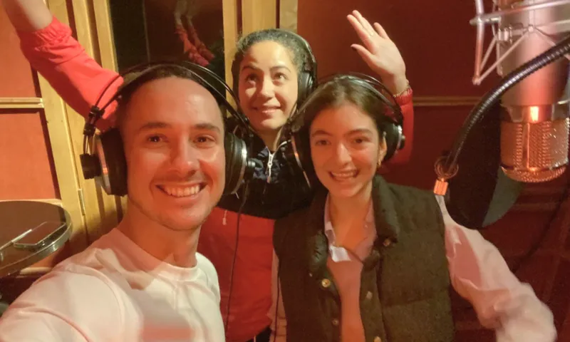 Three people wearing headphones smile and pose for a selfie in a recording studio, with a large microphone visible on the right and padded walls in the background.