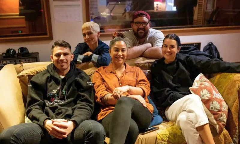 Five people are sitting closely on a couch in a cozy room, smiling at the camera. There is a mix of casual clothing, with some pillows and a blanket visible, suggesting a relaxed and friendly atmosphere.
