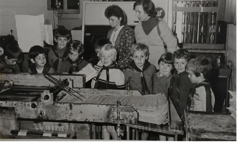 A black and white photo of children visiting a printing press. They are standing around the machines with adults in the background.