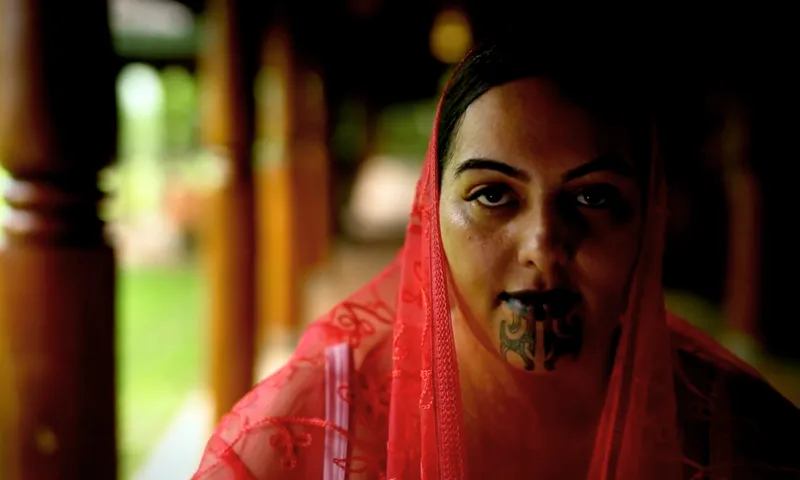 A woman draped in a sheer red veil looks toward the camera, with traditional black chin tattoos visible on her lower face. Wooden columns and greenery are blurred in the background.