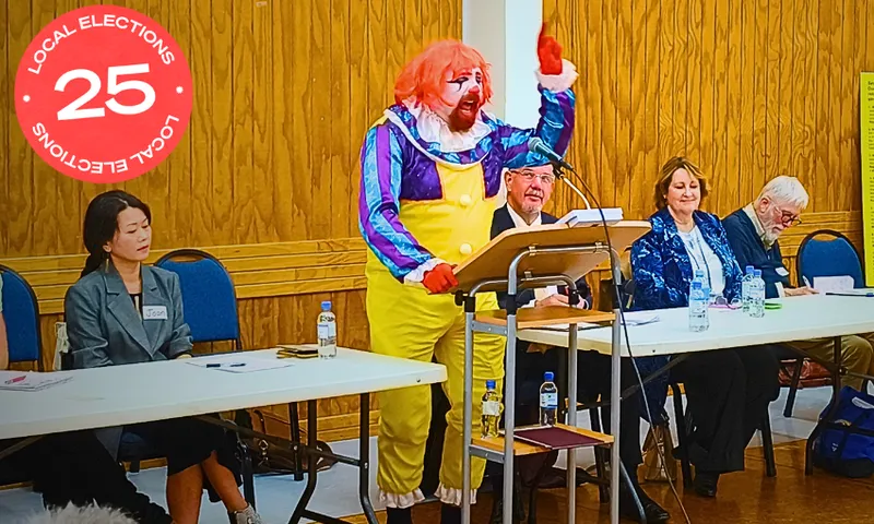 A person dressed as a clown with bright red hair, yellow overalls, and colorful makeup speaks energetically at a podium, while four seated adults in business attire look on in a wood-paneled room.