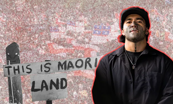 A man with traditional Māori facial tattoos stands in front of a black-and-white sign reading "THIS IS MAORI LAND," with a crowd holding flags in the background.