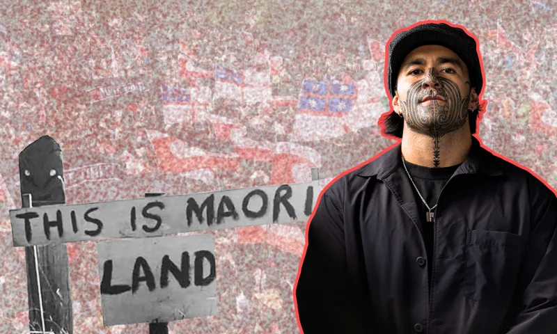 A man with traditional Māori facial tattoos stands in front of a black-and-white sign reading "THIS IS MAORI LAND," with a crowd holding flags in the background.