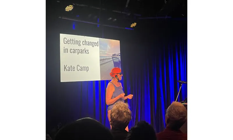 A stage with blue lighting. A screen reads "Getting changed in car parks". There is a woman, Kate Camp, standing in front of the screen wearing togs and a bathing cap.