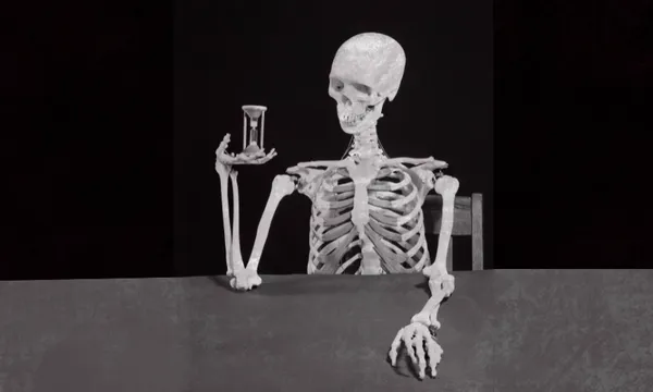 A human skeleton sits at a table, holding an hourglass in one hand and resting its other arm on the table, against a dark background.