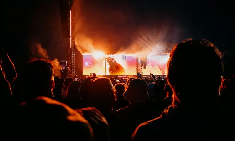 A crowd watches a brightly lit stage at night during a concert or music festival, with orange lights illuminating the performers and some people holding up their phones to record.
