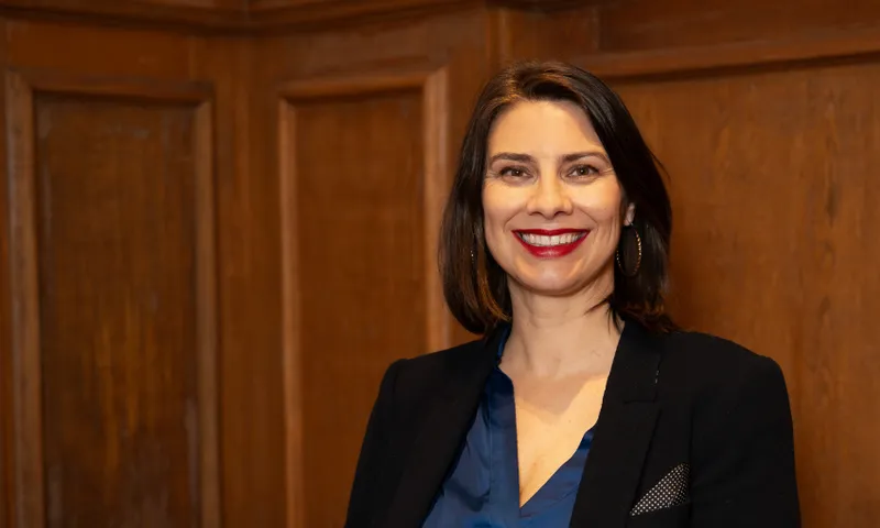 A woman with dark shoulder-length hair and wearing red lipstick smiles to the camera in front of a wooden panelled wall