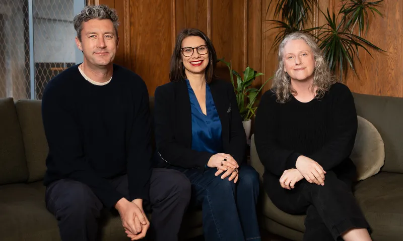 three people smiling while sitting on a couch and wearing dark clothing. one is a man with salt and pepper hair, one is a woman with dark glasses and dark hair and the other is a woman with curly grey hair