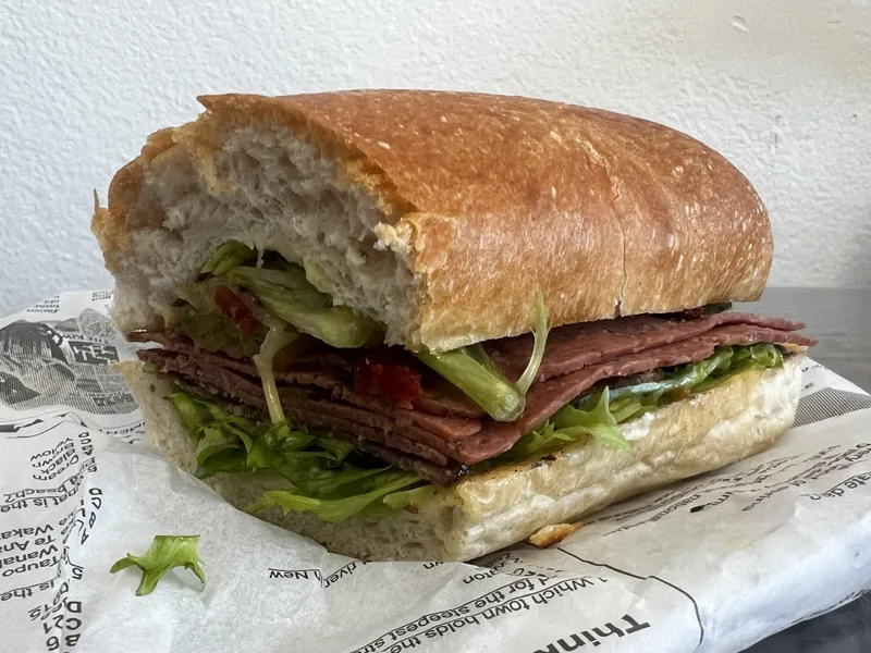 A large sandwich on a hoagie roll with layers of sliced deli meat, lettuce, and tomato, resting on paper with printed text, against a plain light-colored background.