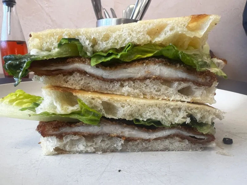 A close-up of a sandwich cut in half, stacked, and filled with breaded meat, lettuce, and sauce on airy focaccia bread, placed on a white plate with utensils and a bottle in the blurred background.