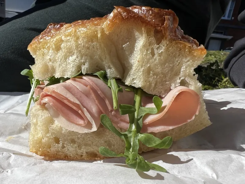 A close-up of a sandwich with fluffy bread, slices of deli meat, and fresh arugula, resting on white paper outdoors in bright sunlight.