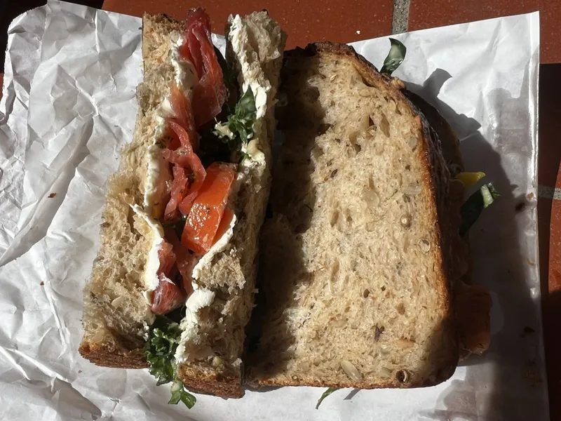 A sandwich made with seeded whole grain bread, filled with cream cheese, leafy greens, tomato slices, and possibly smoked salmon, sits on crumpled white paper on a reddish-brown surface.