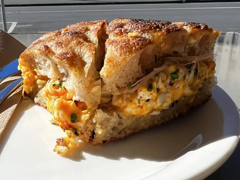 A close-up of a thick sandwich on rustic bread, filled with scrambled eggs, cheese, chives, and turkey, served on a white plate with sunlight highlighting its texture.