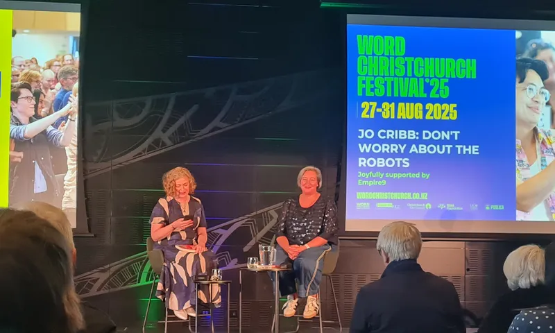 A photograph of an event at Word Christchurch with two women on a dark stage with a large screen behind them. The woman on the left is Susie Ferguson who is wearing a dress and has sort red hair. The woman on the right is Jo Cribb who is wearing pants and a top and has short brown hair.