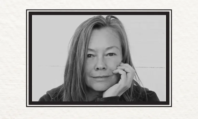 A black and white photo of Kelly Ana Morey who has shoulder length hair and has her hand to her face. She is looking at the camera.