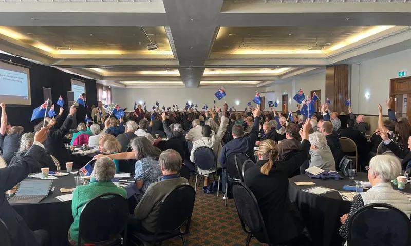 A conference room filled with people sat at tables raise miniature New Zealand flags to raise a vote.