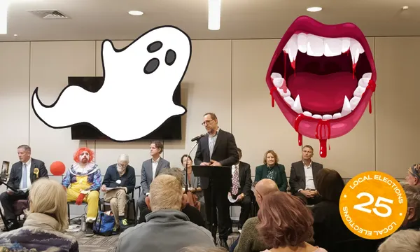 A man speaks at a podium in a crowded meeting room. Seated behind him are people, including someone dressed as a clown. Large cartoon ghost and vampire mouth graphics are added, along with a “Local Elections 25” sticker.