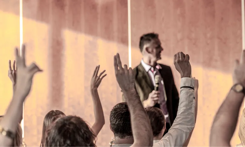 a man in a suit and ties speaks at the front of the room. many people in the group have their hands raised, voting