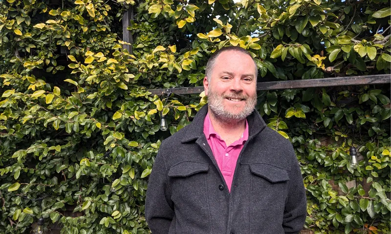 a middle aged man, bald, smiling, pink shirt black jacket, with a bush behind him