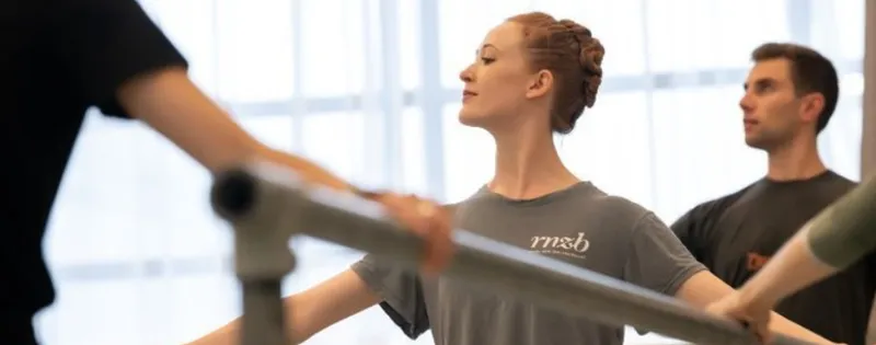 Three dancers are at a ballet barre. The image focuses on a young woman who has her hands spread and she is looking away from the camera.