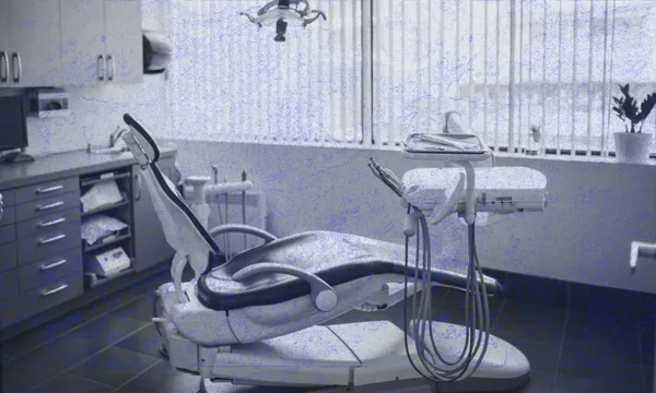 A modern dental office with an empty dental chair, dental tools, and equipment. Cabinets and a computer are on the left, and a window with vertical blinds lets in natural light. A potted plant sits on the right.