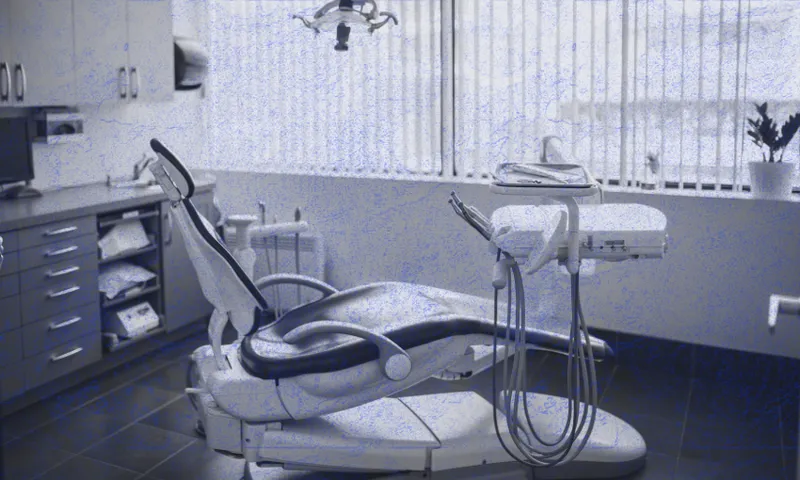 A modern dental office with an empty dental chair, dental tools, and equipment. Cabinets and a computer are on the left, and a window with vertical blinds lets in natural light. A potted plant sits on the right.