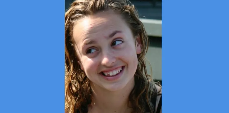 A young person with curly, wet hair smiles and looks to the side against a blurred outdoor background. The image has blue borders on the left and right sides.