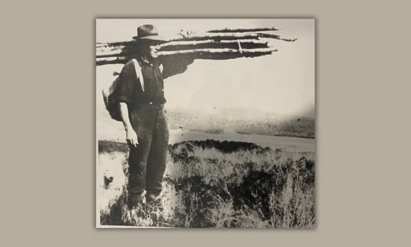 a man in a work shirt, pants, boots and cowboy hat carries long sticks