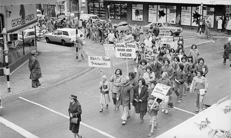 A black-and-white photo shows a group of people marching down a city street, holding signs demanding equality for Māori language and culture. Bystanders watch, and a police officer stands at the corner.