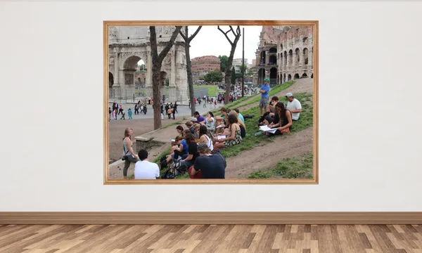 A group of people sit and write on a grassy slope near the Colosseum in Rome, seen through a large framed picture hanging on a plain white wall in a gallery with a wooden floor.