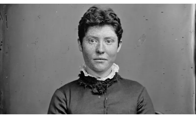 A black and white photo of a historic woman (19th century) wearing a high-necked dress with a collar; and short hair. She has freckles all over her face and is staring into the camera lens.