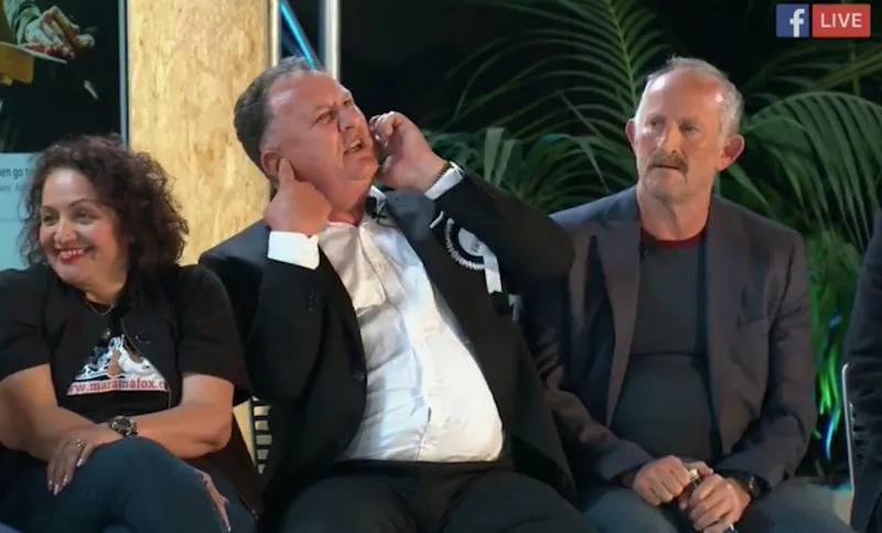 Three people sit side by side; the man in the center covers one ear while talking on the phone, looking distressed, while the woman on the left smiles and the man on the right looks on calmly. A Facebook Live icon appears in the corner.