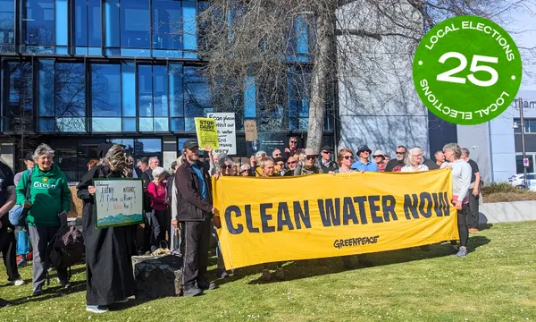 A group of people holding a large banner that says "Clean Water Now" in the sunshine