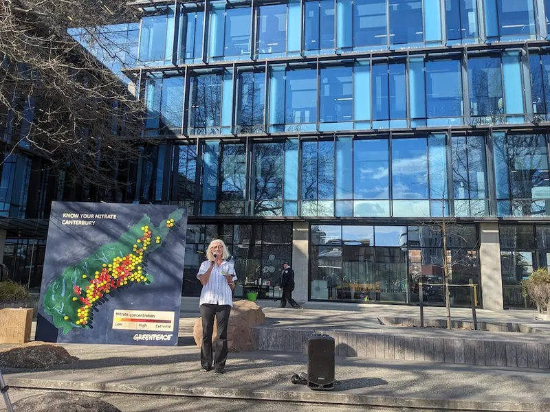 Kate Gillard, a woman with white hair in a white shirt, holds a water bottle. in the background is a quite wonky map of the south island with clusters of red and purple indicating high nitrate levels, and the glassy facade of the Environment Canterbury building
