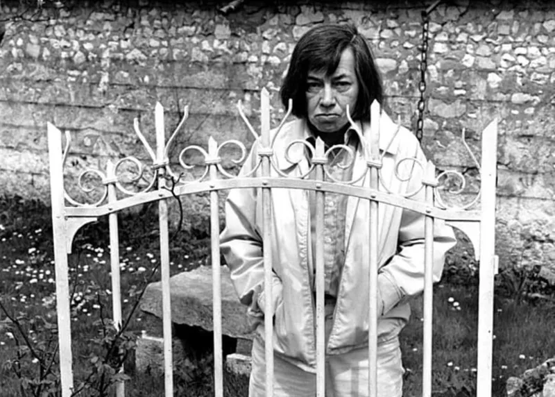 Patricia Highsmith stands behind a wrought iron gate, glaring in the camera, her hands in the pockets of her jacket.