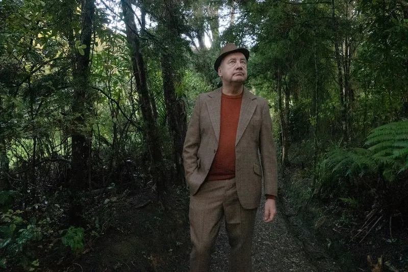 A man wearing a brown suit, matching hat, and orange sweater walks along a wooded forest path, surrounded by green trees and foliage.