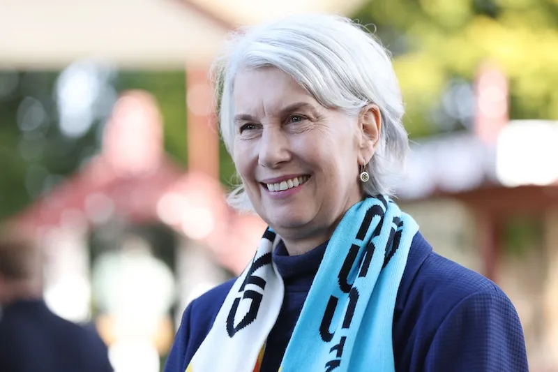 A smiling older woman with short gray hair, wearing drop earrings and a blue scarf with black and yellow patterns, stands outdoors with blurred greenery and structures in the background.