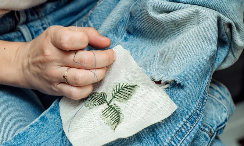 A photo showing a hand mending torn jeans, using a floral motif.