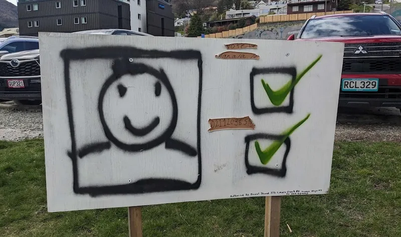 A wooden sign displays a spray-painted smiley face next to two checkboxes, both marked with green checkmarks. It stands outdoors on grass, with parked cars and buildings in the background.
