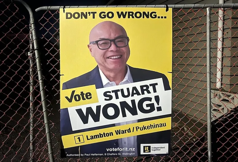 A campaign poster on a fence features a smiling man in glasses and a suit. The text reads: “DON’T GO WRONG… Vote STUART WONG! Lambton Ward / Pukehinau, 1.” The candidate’s website and party logo are also visible.