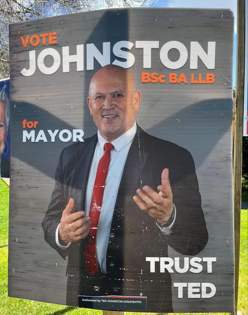 Campaign poster showing a bald man in a suit and red tie, smiling and gesturing with his hands. Text reads: “VOTE JOHNSTON BSc BA LLB for MAYOR. TRUST TED.” The poster is displayed outdoors on a sunny day.