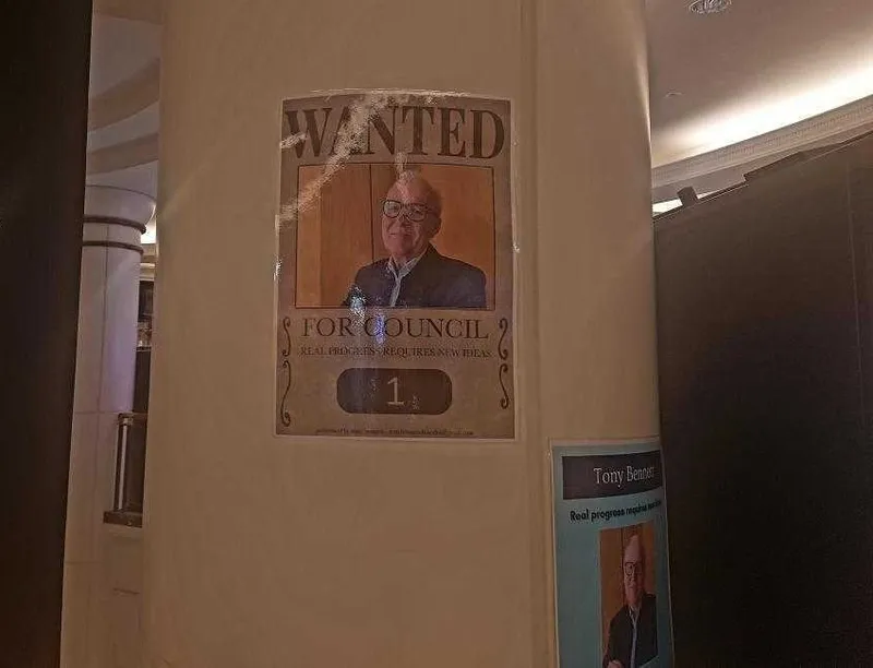 A poster on a beige pillar features an older man in glasses and a suit with the large text "WANTED for Council" above his photo. Another smaller poster with his photo is partially visible on the right.