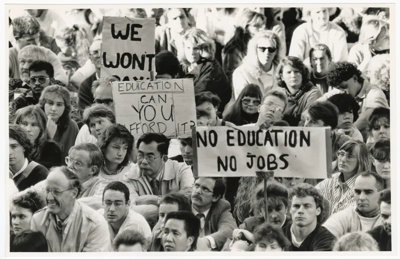 a black and white photo of a crowd of students siting holding placards protesting student fees