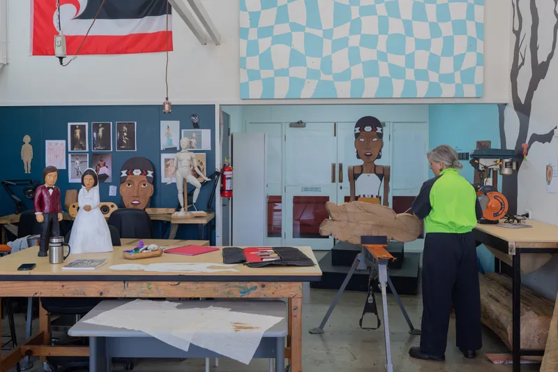 A person in a green shirt works on a wooden sculpture in an art studio filled with tools, wooden figures, drawings, and a large flag on the wall. The workspace is creative and organized.
