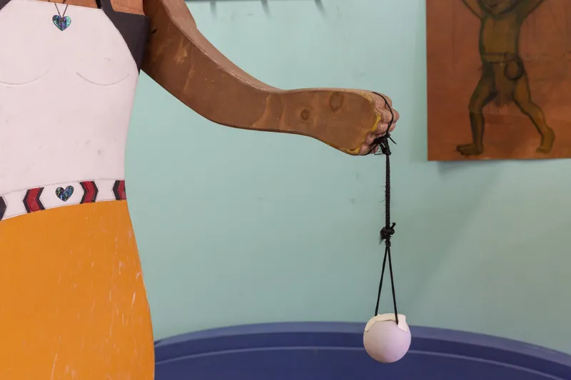 A wooden sculpture of a person in a yellow and white dress holds a string with a white ball attached. The background is light blue with a brown sketch of a figure on the wall.