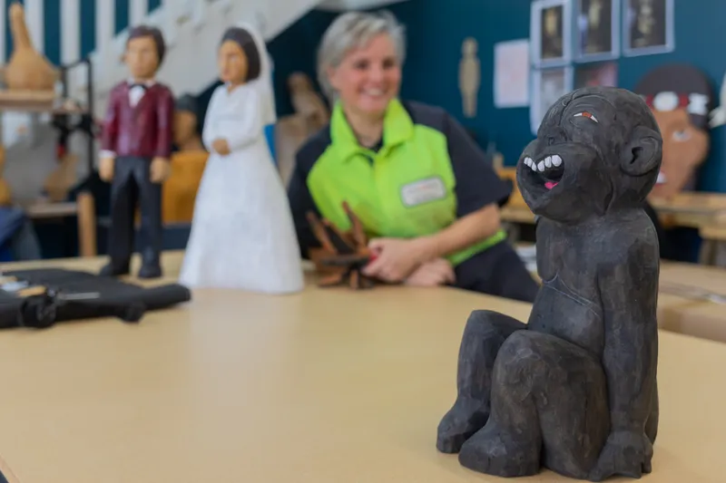 A carved wooden figure of a monkey sits on a table in the foreground, while a smiling person in a green shirt and two wooden human figures are out of focus in the background.